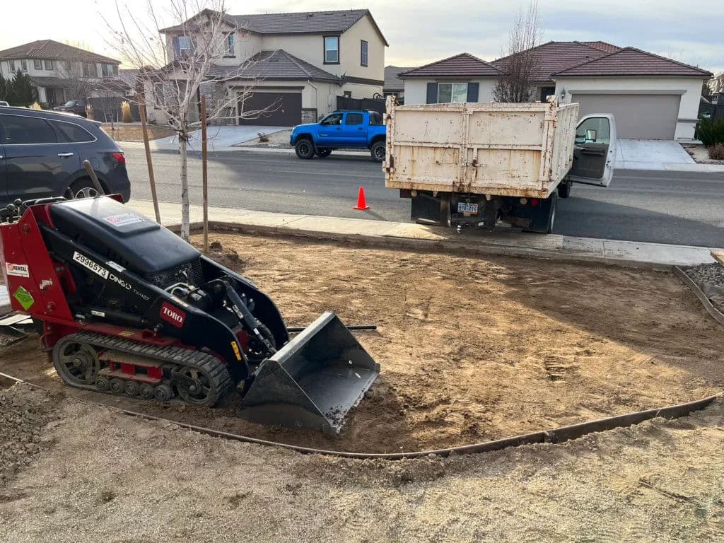 Mini bulldozer on gravel driveway with parked truck and houses in background.