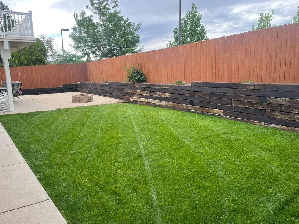 Lush green lawn with wooden fencing and a stone fire pit in a backyard setting.