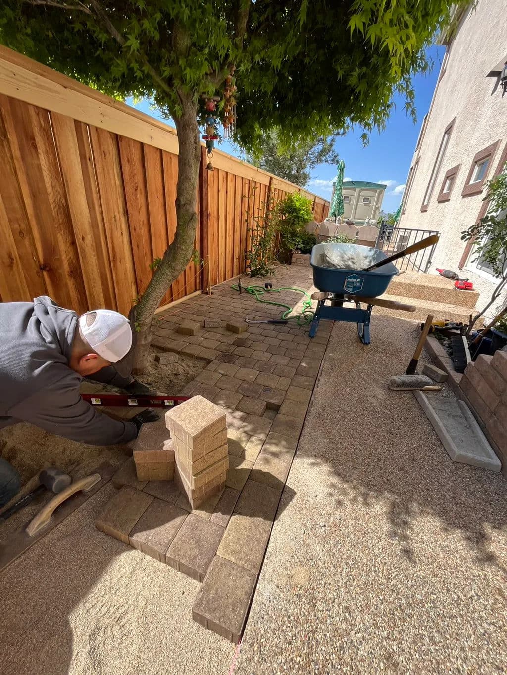 Person laying pavers in a landscaped yard with a wheelbarrow and tools nearby.