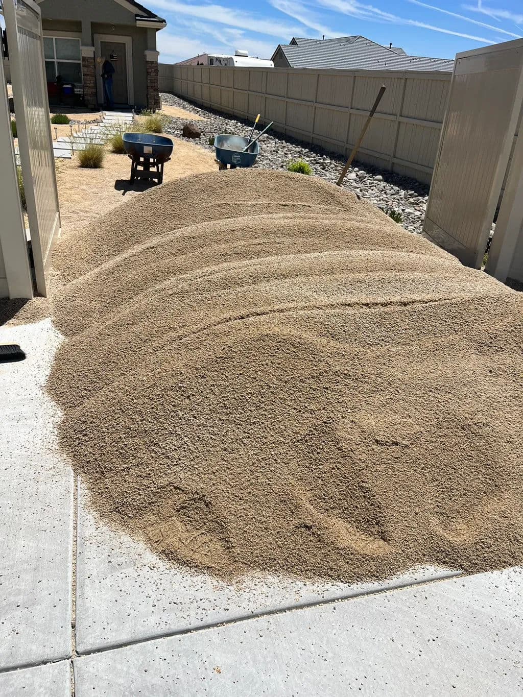 Pile of sand in a residential yard with wheelbarrow and tools in the background.
