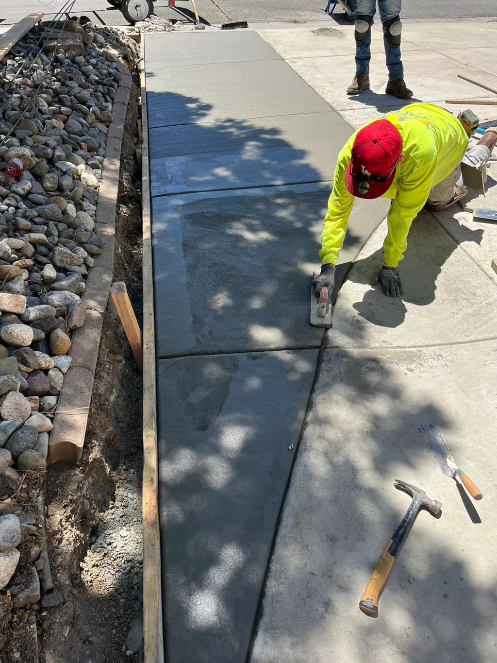 Worker smoothing freshly poured concrete sidewalk with tools on a sunny day.