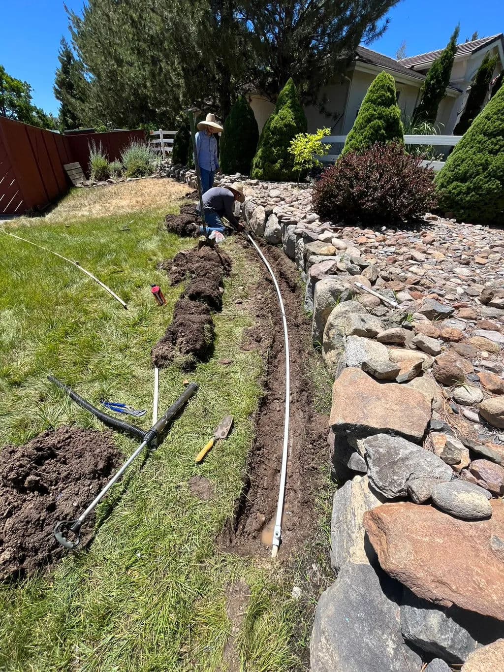 Person digging a trench for irrigation installation in a landscaped yard with rocks and plants.