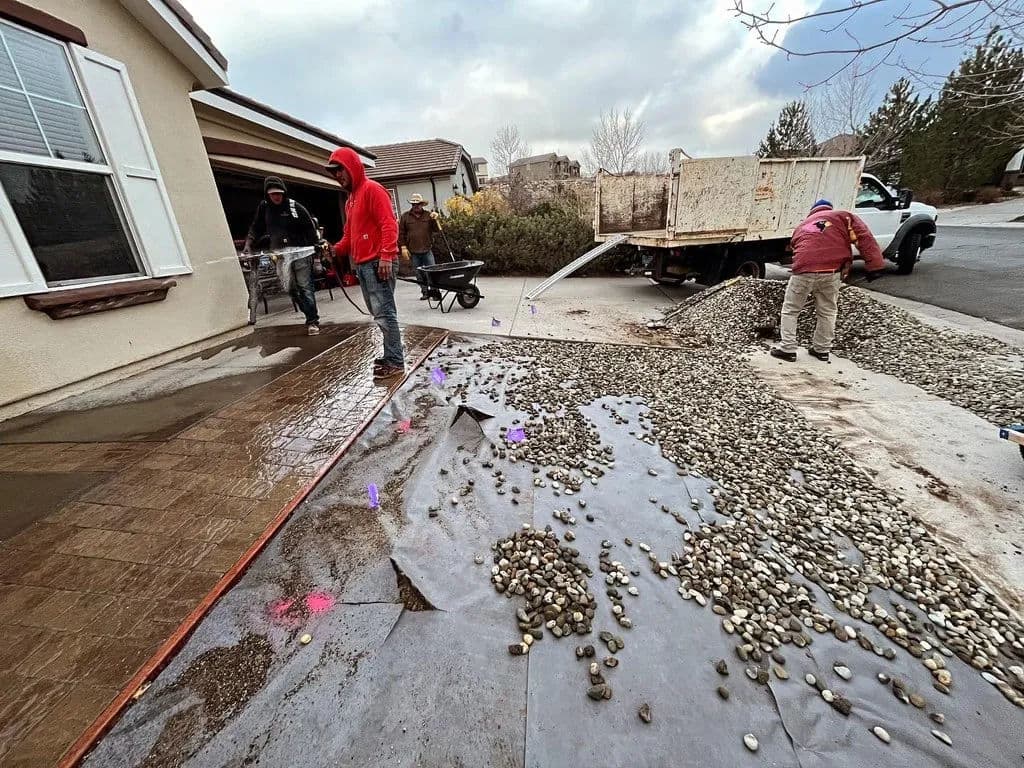 Construction crew pouring concrete on driveway with gravel and equipment nearby.