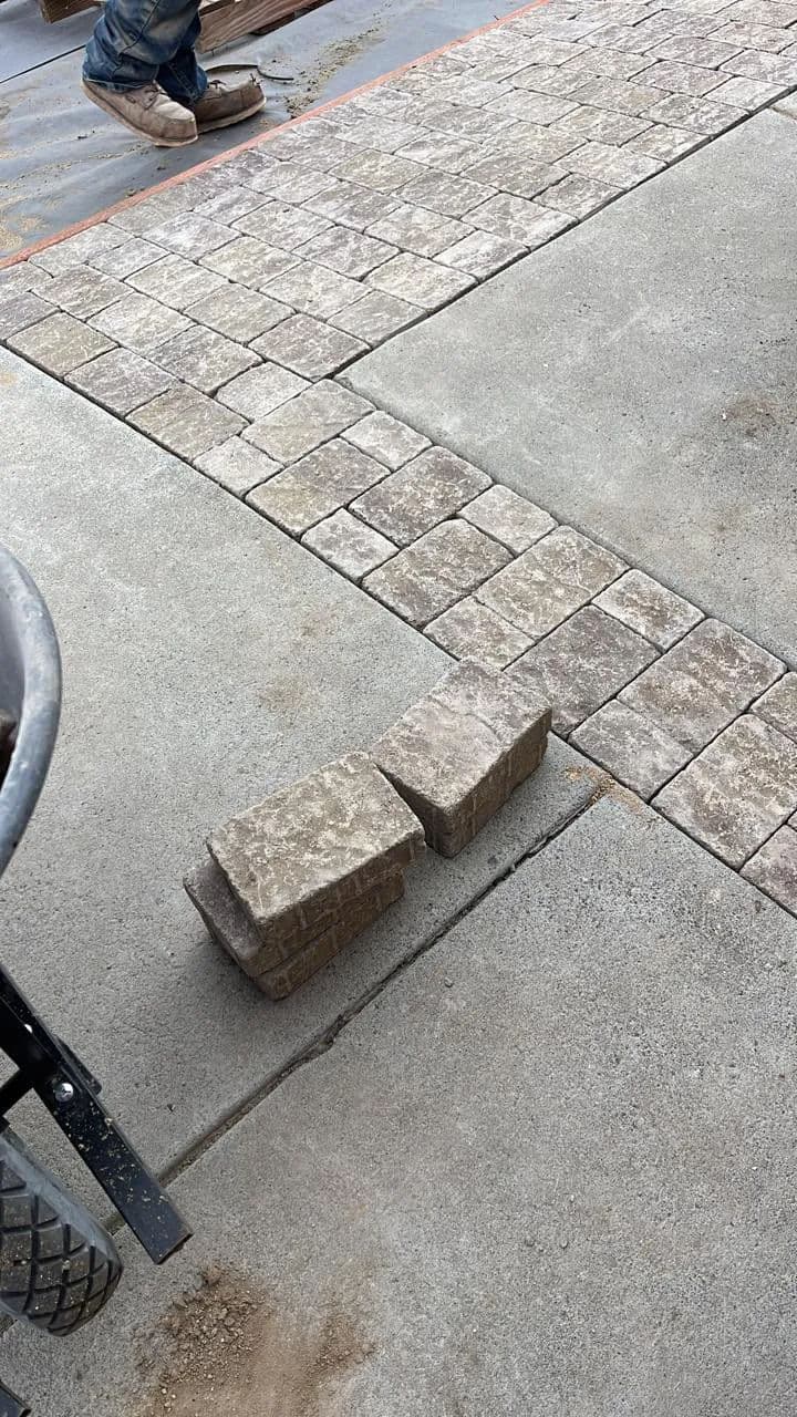 Worker installing stone pavers on a patio, with tools and completed paver section visible.
