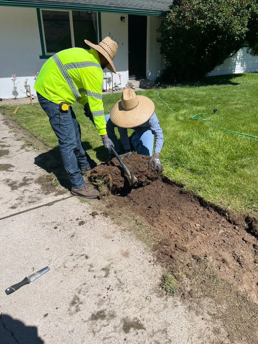 Two workers in straw hats digging a trench alongside a grass lawn.