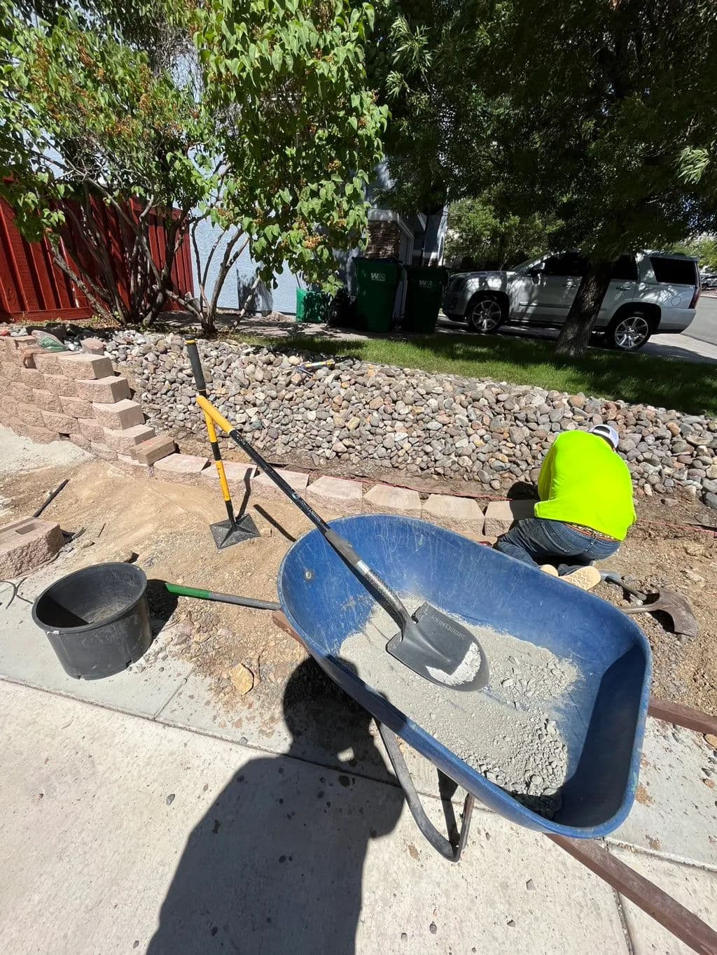 Construction worker using tools to lay concrete in a landscaped area with a wheelbarrow.