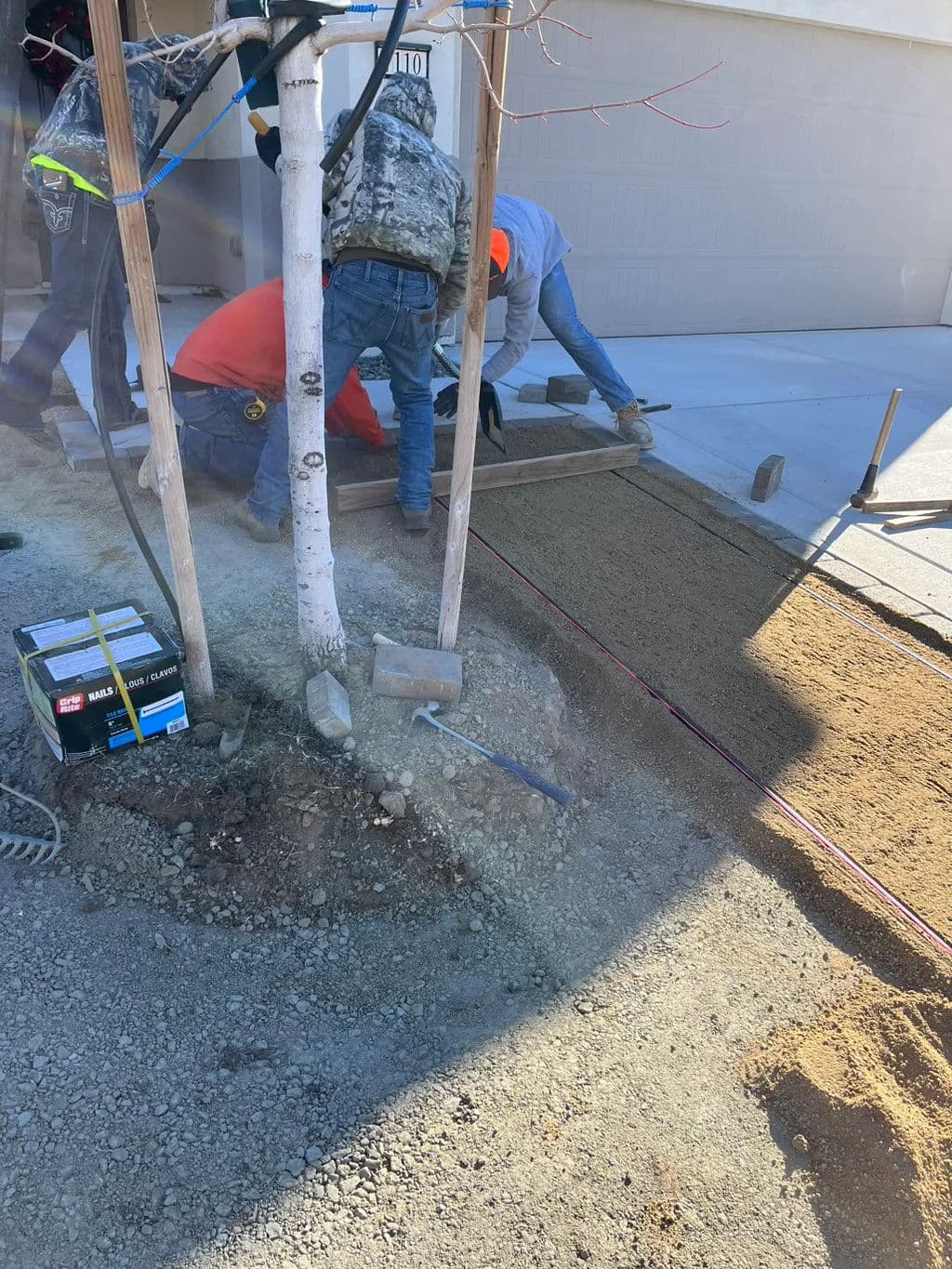 Workers preparing landscape near a garage, leveling soil and using tools around a tree.