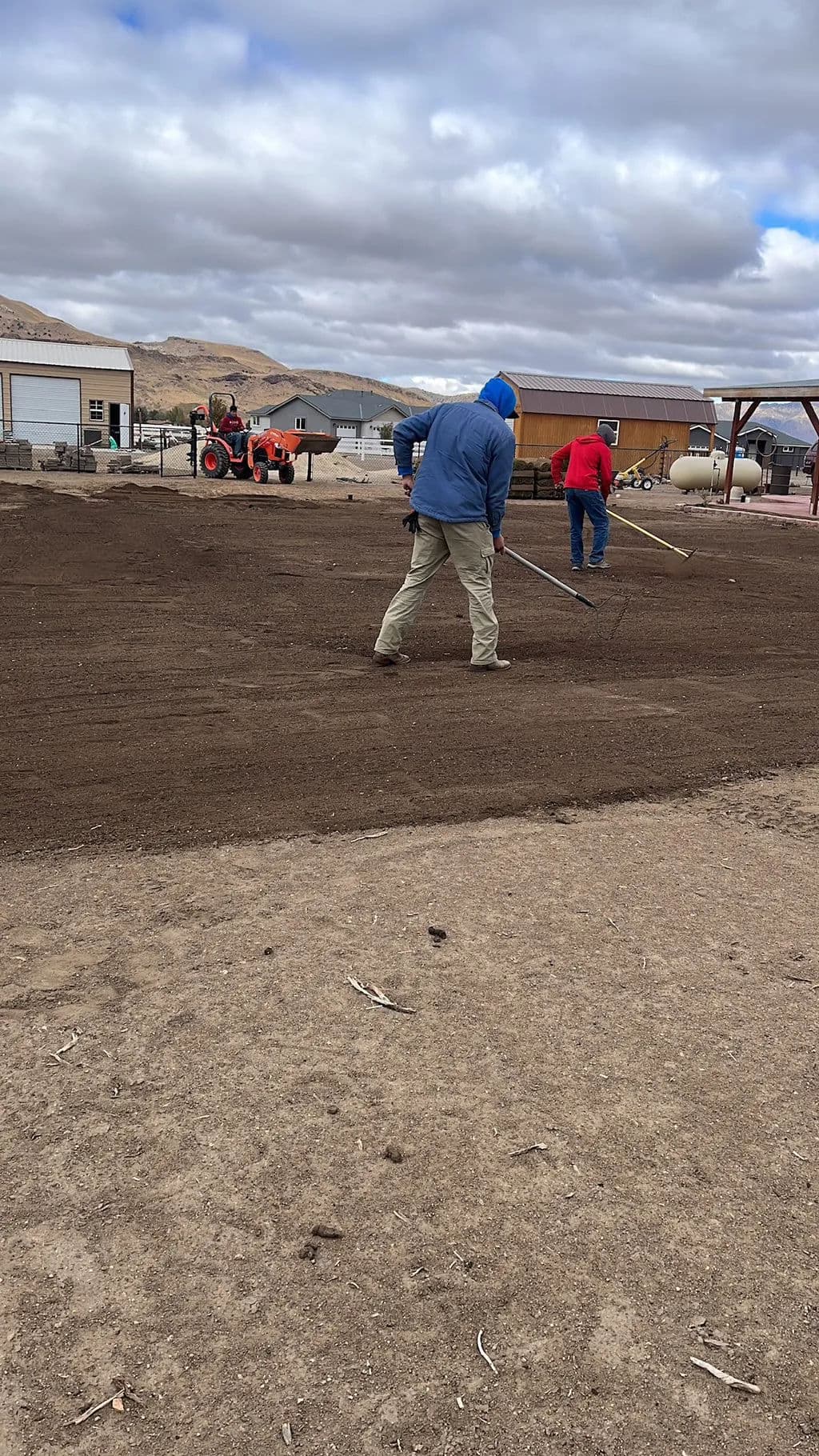Two men leveling soil with tools on a farm with a tractor in the background.