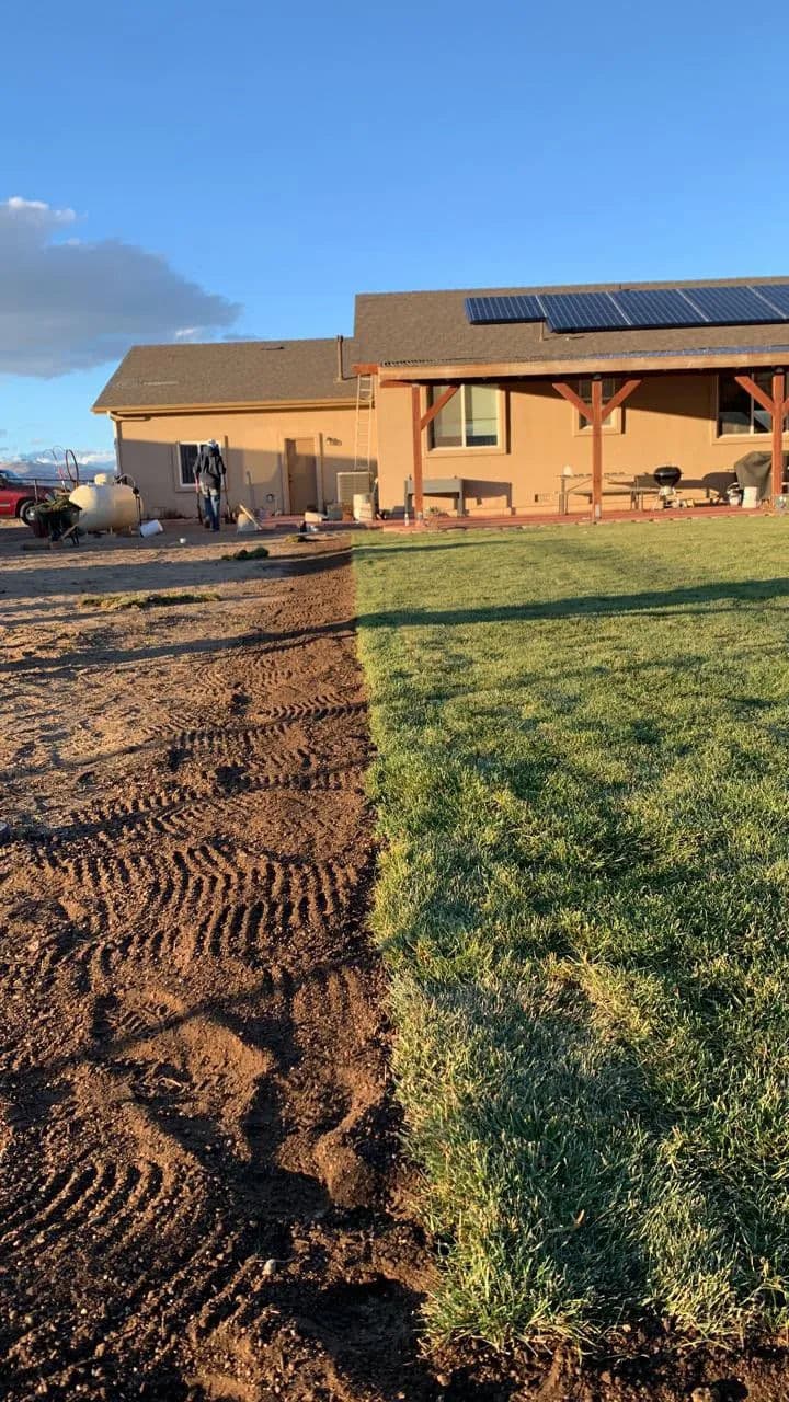 Half freshly tilled soil and half lush green grass beside a home with solar panels.