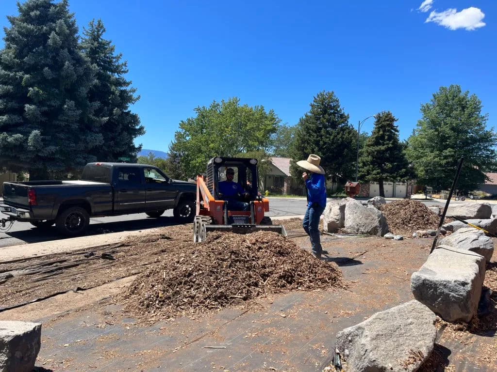 Landscaping crew using machinery to move mulch and rocks in a residential area.