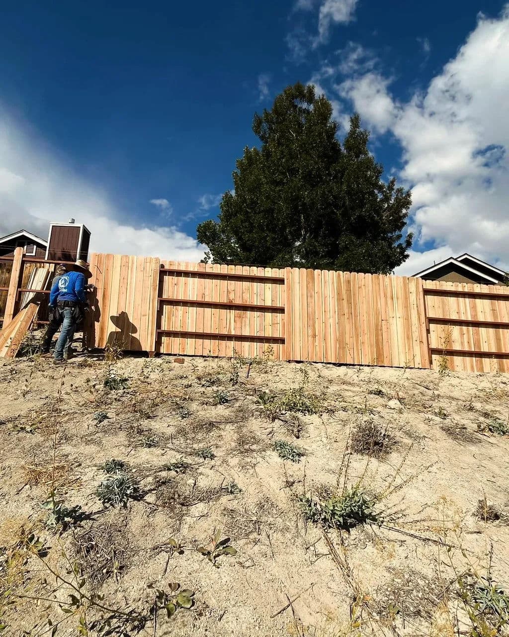 Worker constructing a wooden fence on a hillside under a cloudy sky.