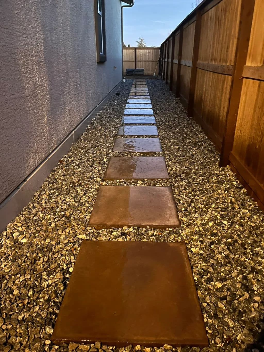 Stone pathway over gravel alongside a wooden fence, leading toward a house.