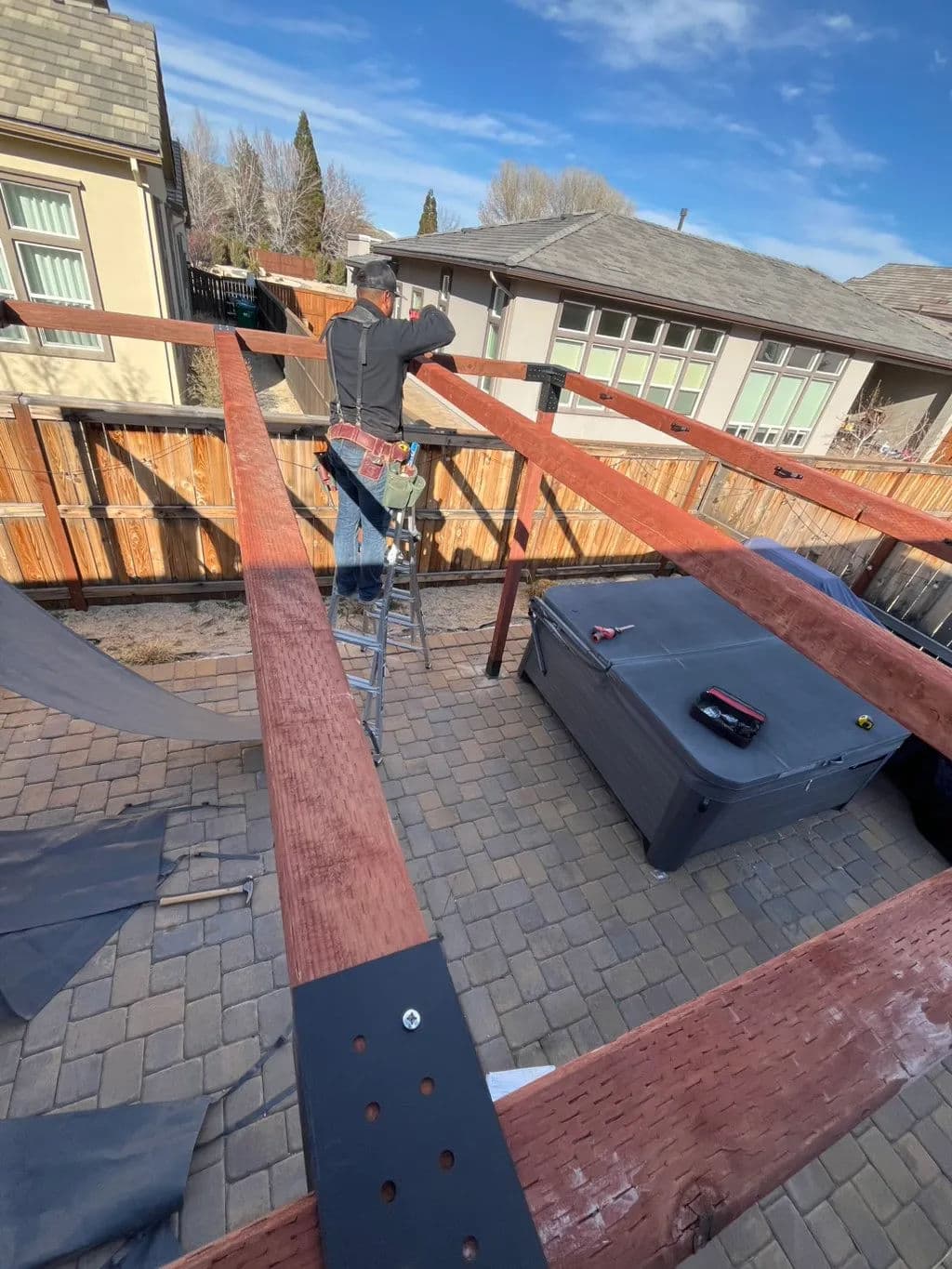 Man constructing a wooden pergola in a backyard with a hot tub and blue sky backdrop.