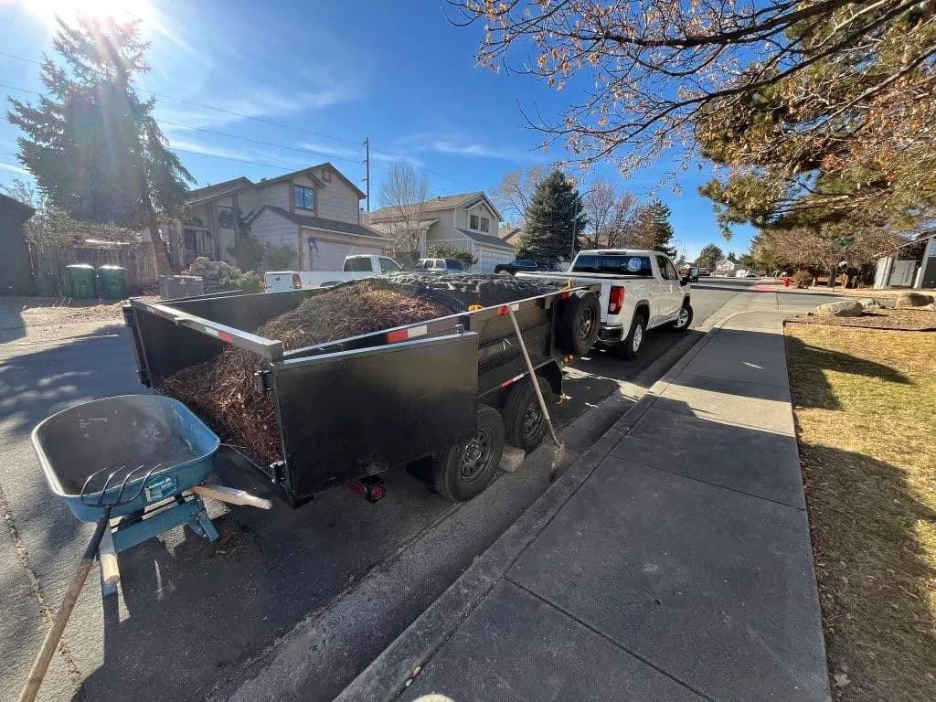 Trailer filled with leaves parked on the street beside a pickup truck and wheelbarrow.