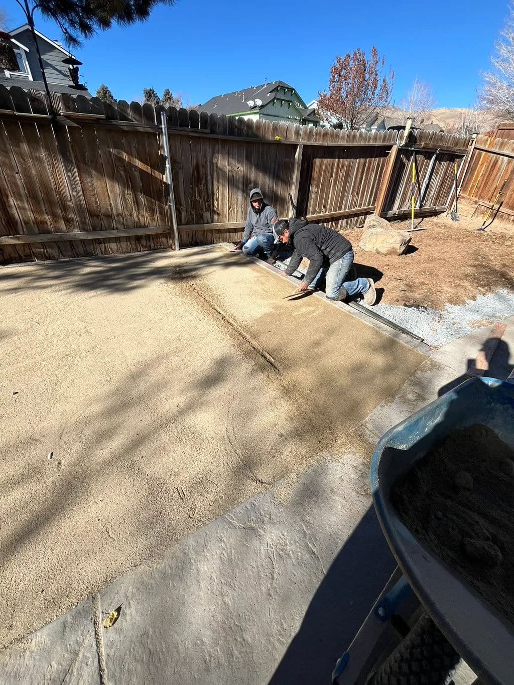 Workers leveling sand for a patio installation in a residential backyard under blue sky.