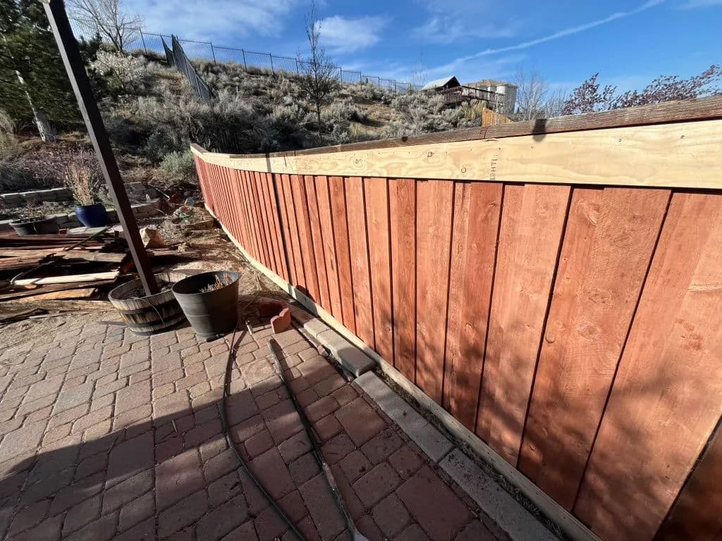 Newly built wooden fence curving along a garden patio with clear blue sky in the background.