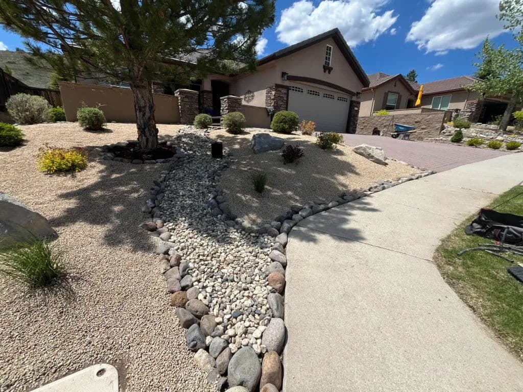 Landscaped front yard with gravel pathway, shrubs, and a tree near the house under a blue sky.